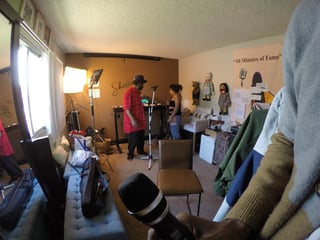 Indoor behind-the-scenes look at a film or TV production set with crew members, lighting equipment, clothing racks, and props scattered around the room