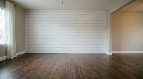 Empty room with white shiplap accent wall, dark hardwood flooring, and large window with white curtains