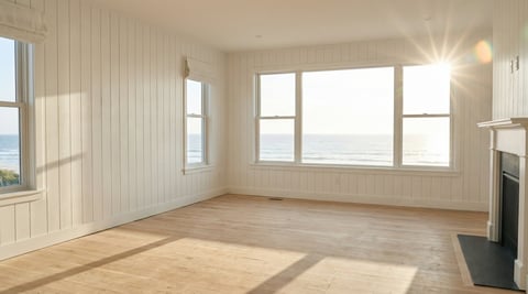 Empty bright room with hardwood floor, white beadboard walls, large windows with ocean view, and fireplace