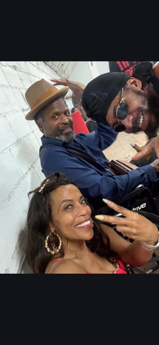 Group of people posing together for a selfie indoors, with one woman smiling at the camera in the foreground and men behind her wearing hats and casual attire