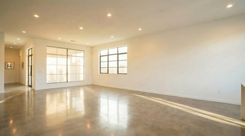 Empty modern living room with polished concrete floors, large windows with natural light, recessed ceiling lights, and white walls