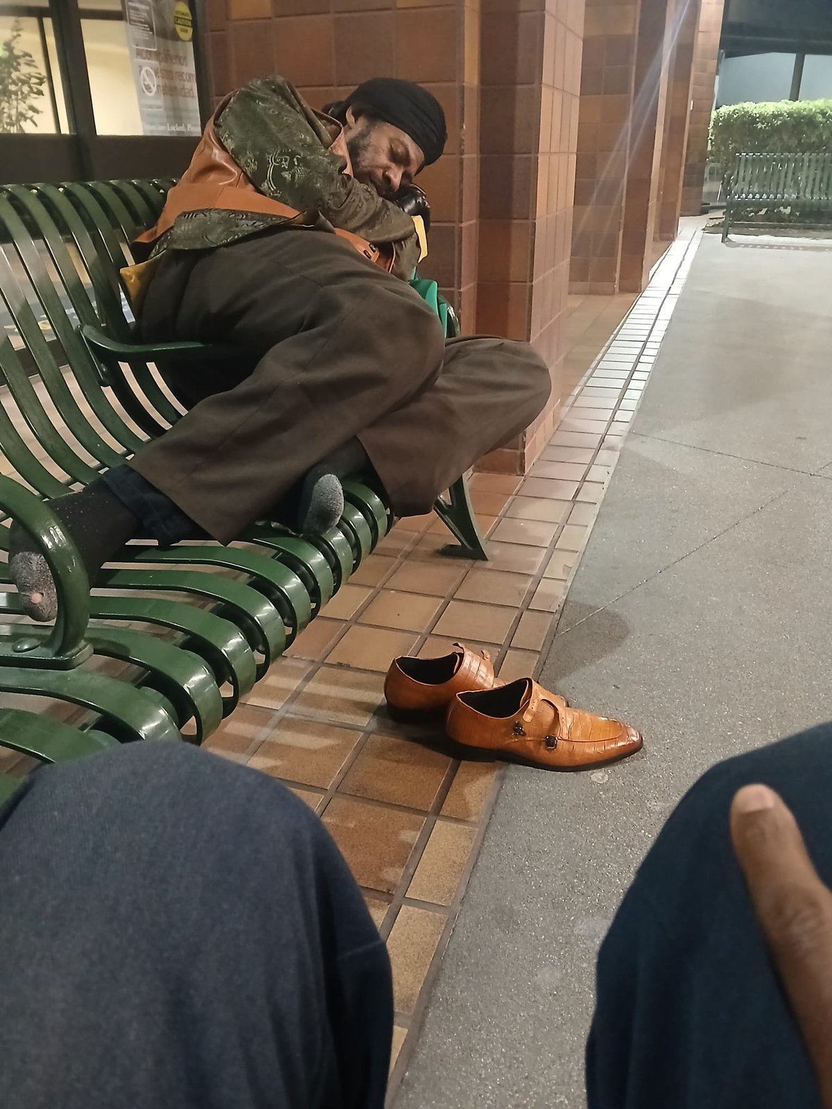 Person sitting on a green bench holding a lizard, wearing brown shoes, on a tiled outdoor patio area