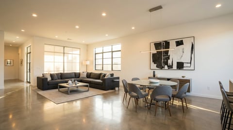 Modern open-concept living room with gray sectional sofa, dining table, concrete floors, and abundant natural light from large windows
